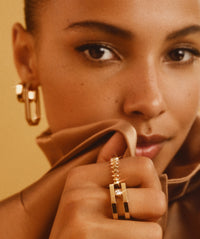 A close-up of a model wearing a MIRACO Geometric Double Band and Solitaire Gold Diamond Ring and gold earrings set against a warm beige background.
